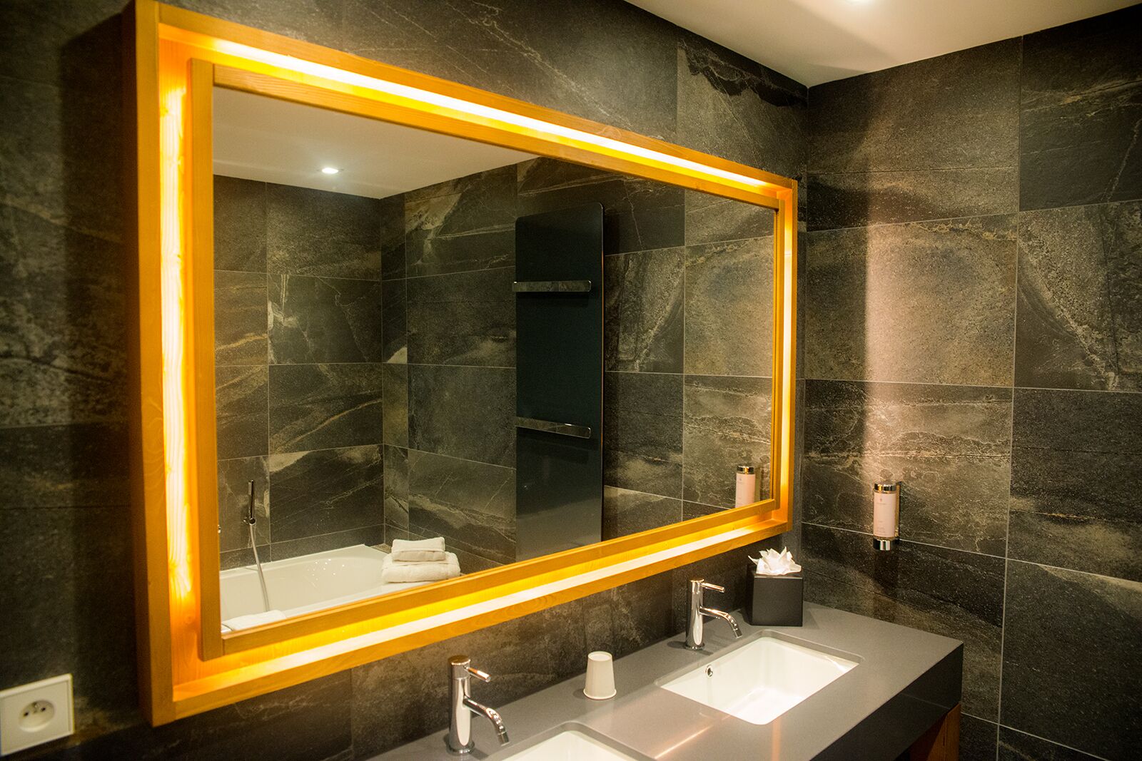 Un baño moderno con baldosas de mármol oscuro, un espejo grande con marco dorado, dos lavamanos y un perchero para toallas.