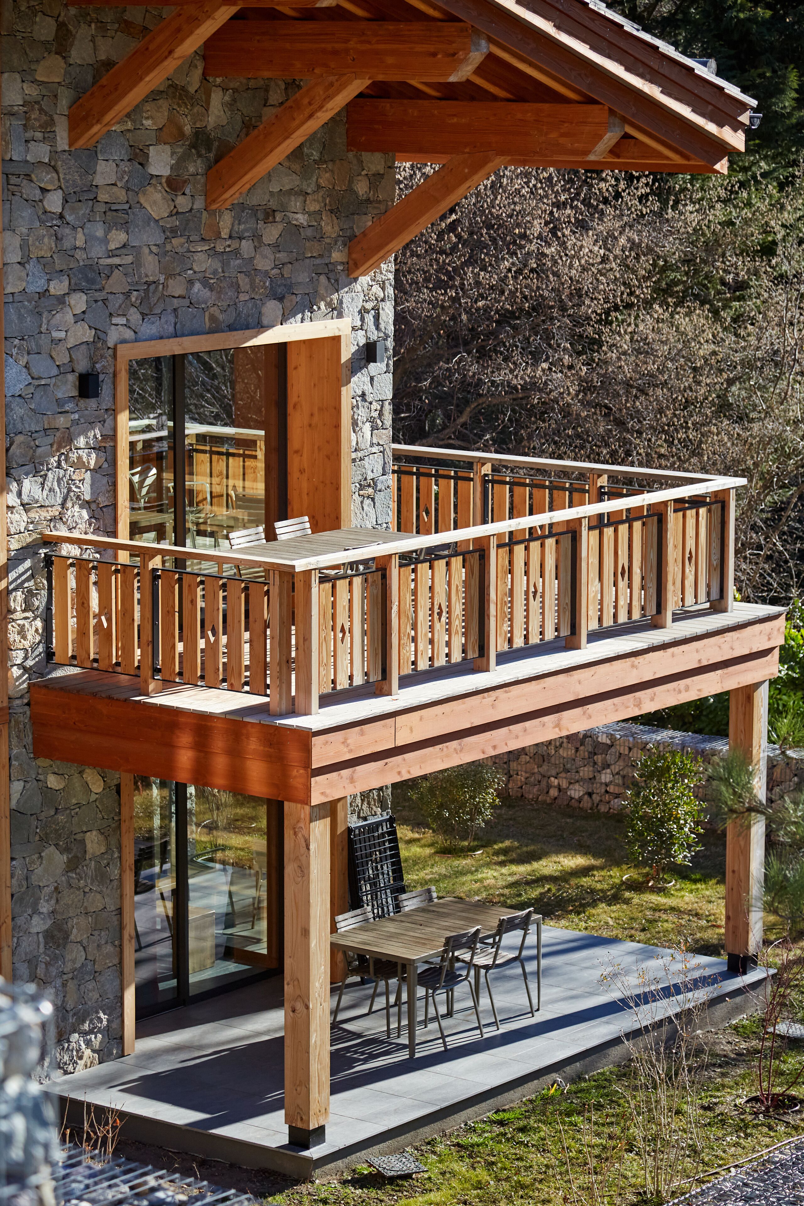 A wooden deck with a railing, a stone wall, and a glass door leading to a patio area with a table and chairs.