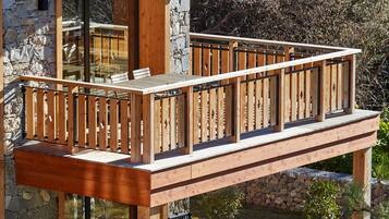 A wooden deck with a railing, a stone wall, and a glass door leading to a patio area with a table and chairs.