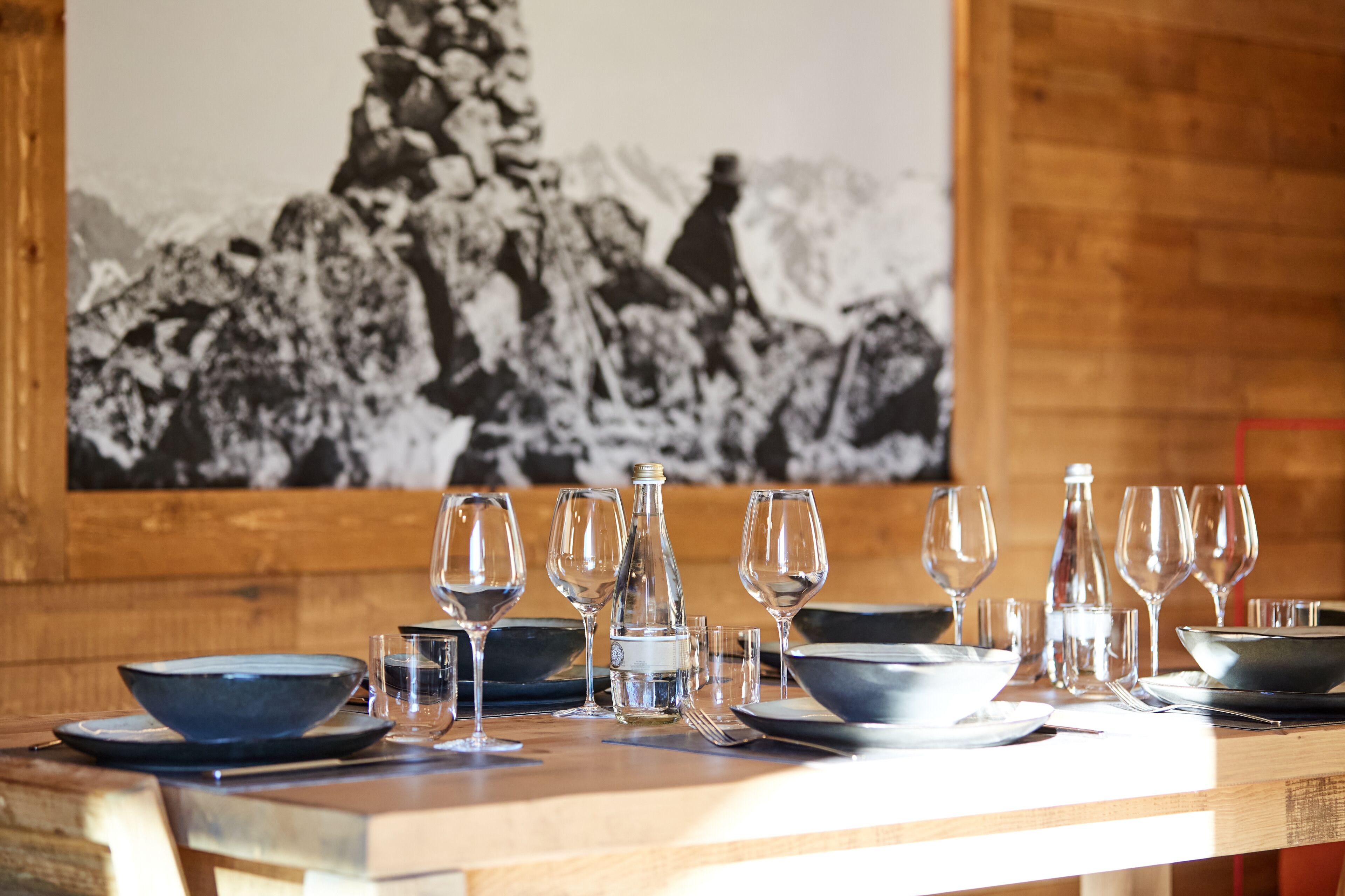 A dining table set with clear glassware and plates, a bottle, and a bowl, with a mountainous landscape in the background.