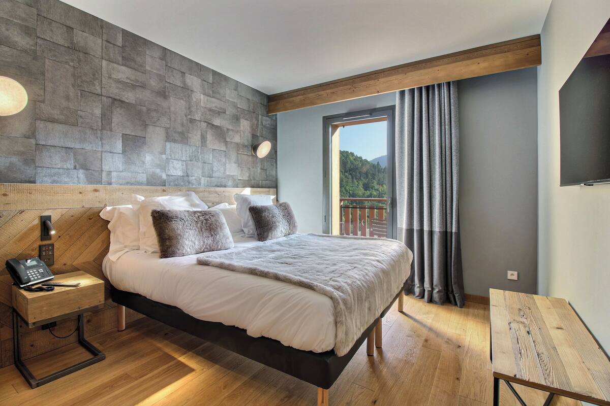 A modern bedroom with a wooden headboard, a large bed with white bedding, a bedside table with a phone, and a view of a balcony with curtains.