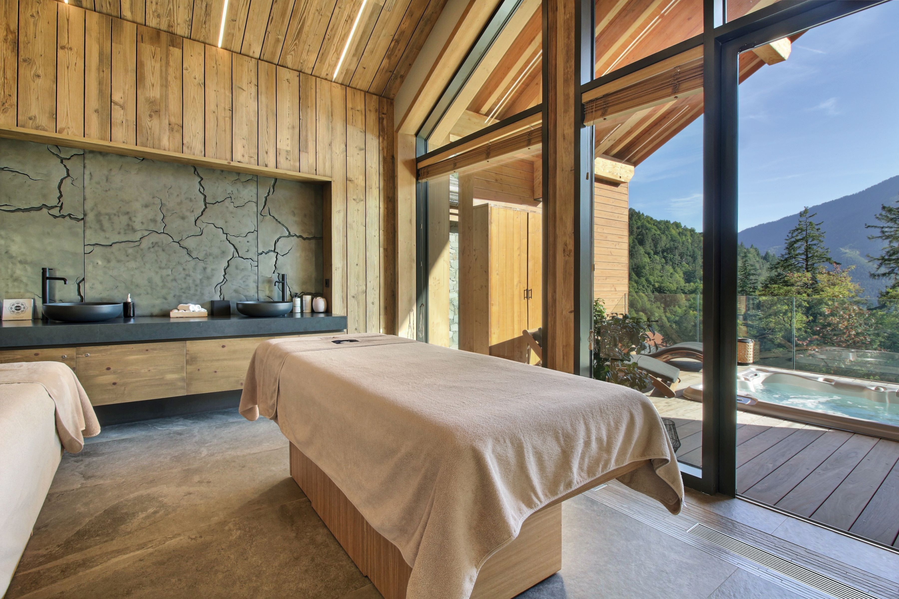 A spa room with a massage table, wooden paneling, a stone wall, and a view of a hot tub and mountains.