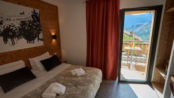 A hotel room with a bed, a wooden headboard, a large poster, a red curtain, and a balcony with a view of mountains.