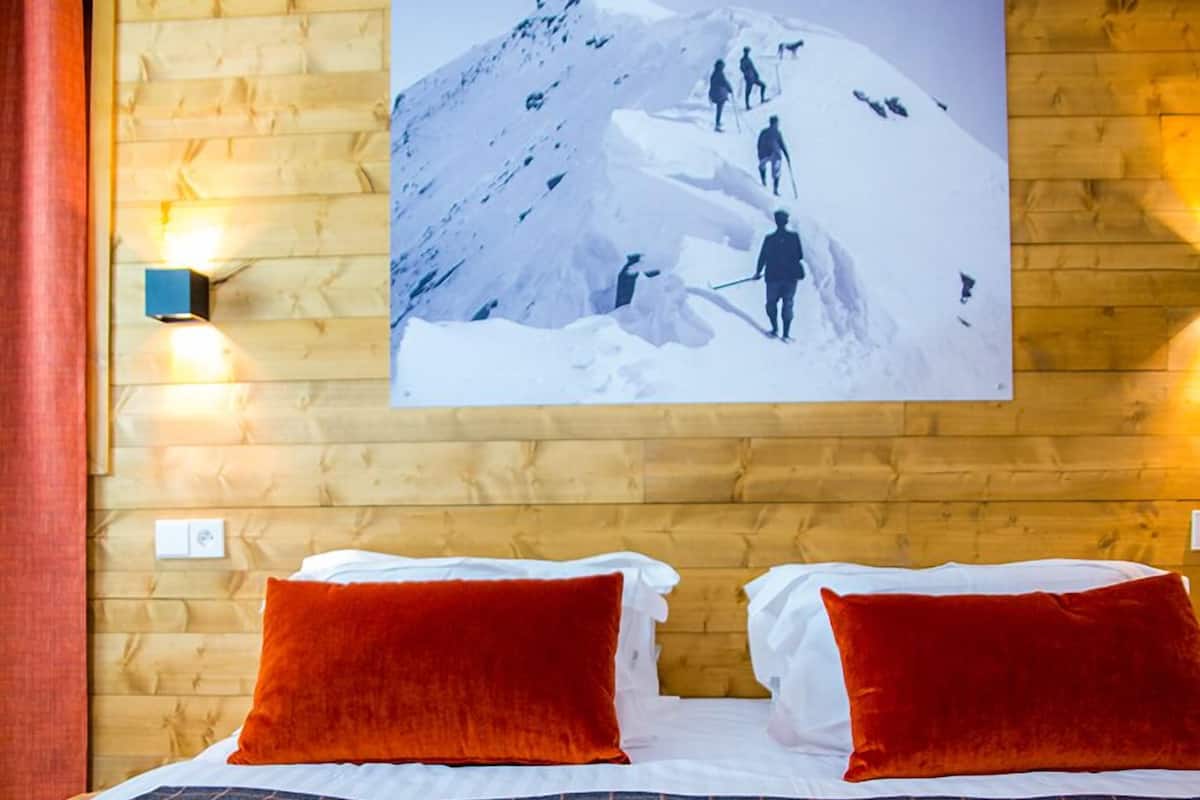A hotel room with a wooden wall, a large bed with a checkered bedspread, two red pillows, and a framed picture of climbers on a mountain.
