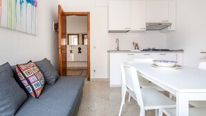 Une cuisine moderne avec des meubles blancs, une table à manger blanche et un canapé gris agrémenté d’un coussin coloré.