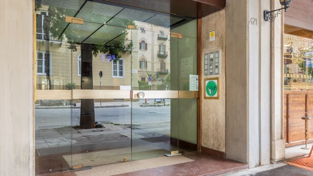 Une entrée d’immeuble avec une porte en verre, donnant sur la rue, et un panneau indiquant une zone où le stationnement est interdit.