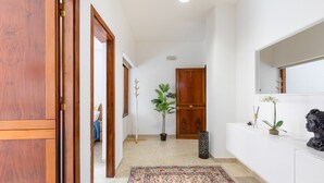 Un couloir avec un tapis à motifs, des portes en bois, une plante en pot, une petite poubelle et un buste posé sur un comptoir blanc.