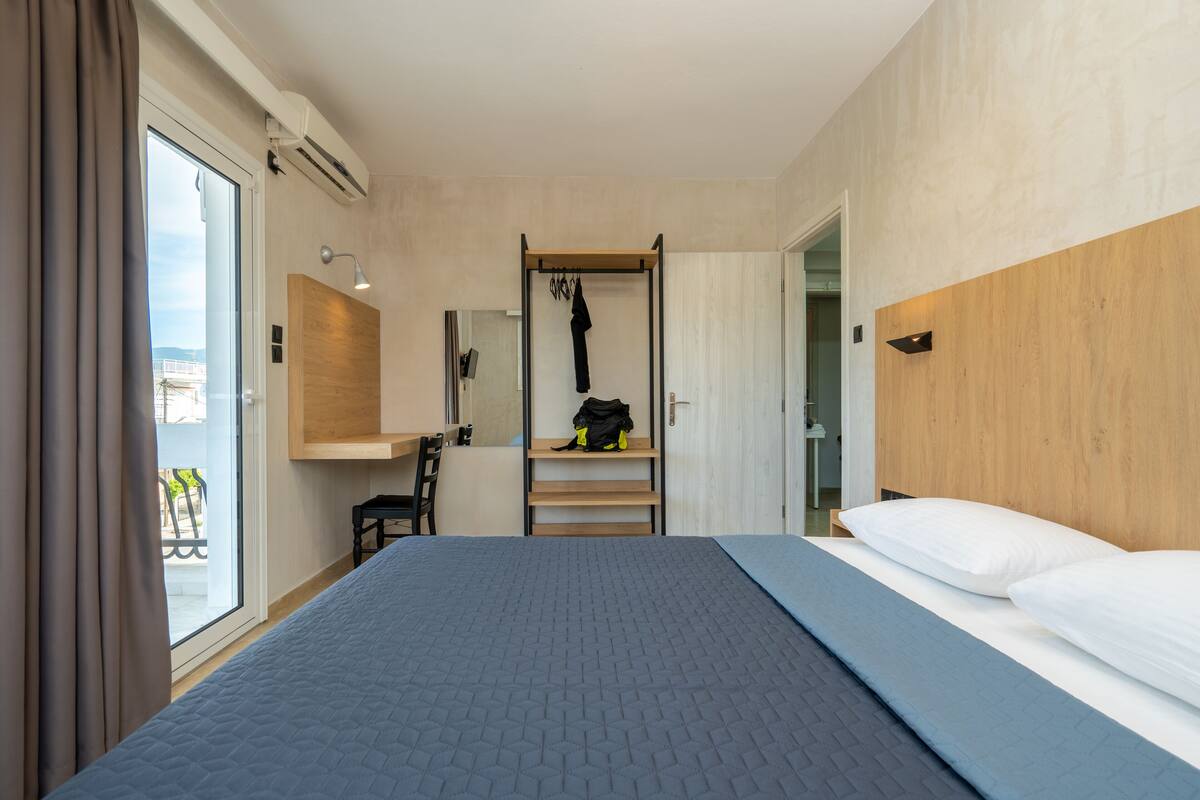 Quarto de hotel moderno com uma cama grande, cabeceira de madeira, prateleira embutida e porta deslizante para uma varanda.