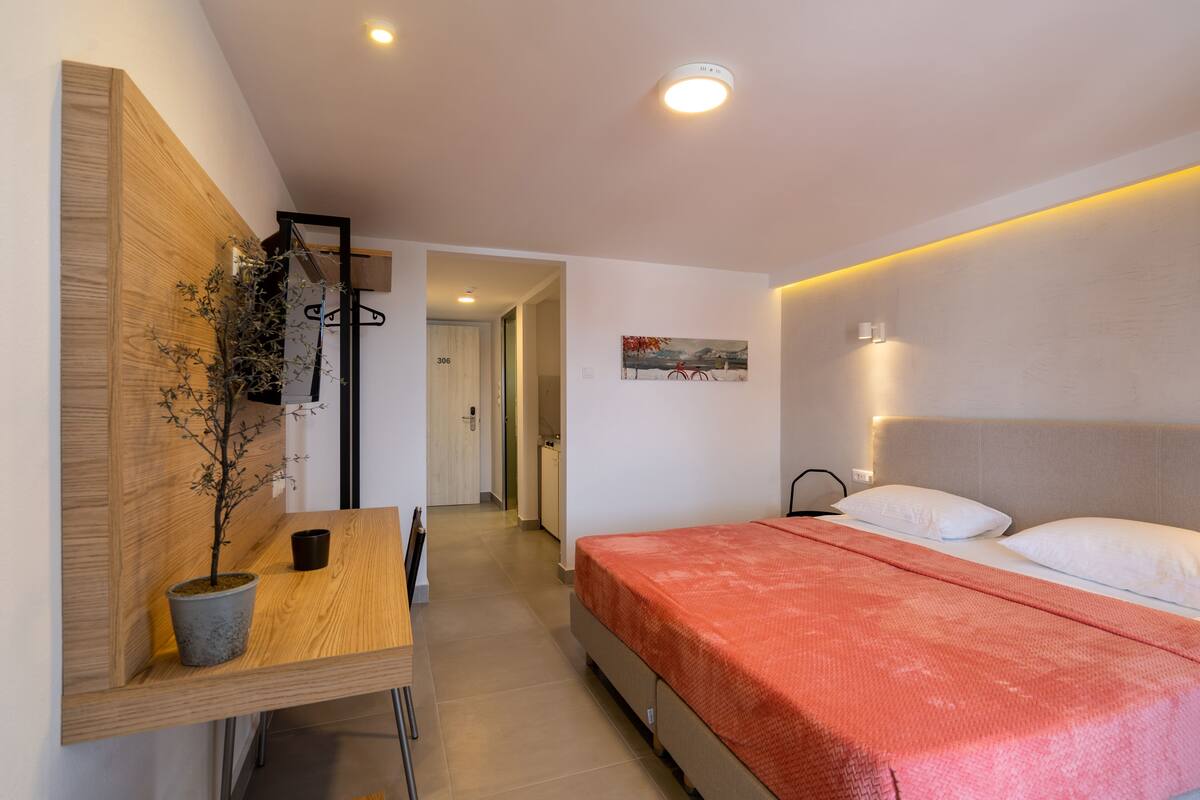 Quarto moderno com cabeceira de madeira, cama com manta vermelha, mesa de cabeceira com planta em vaso e corredor que leva a outro cômodo.