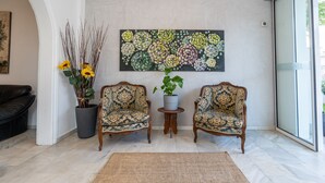 A living room with two patterned armchairs, a coffee table, a large floral painting, and a potted plant with yellow flowers.