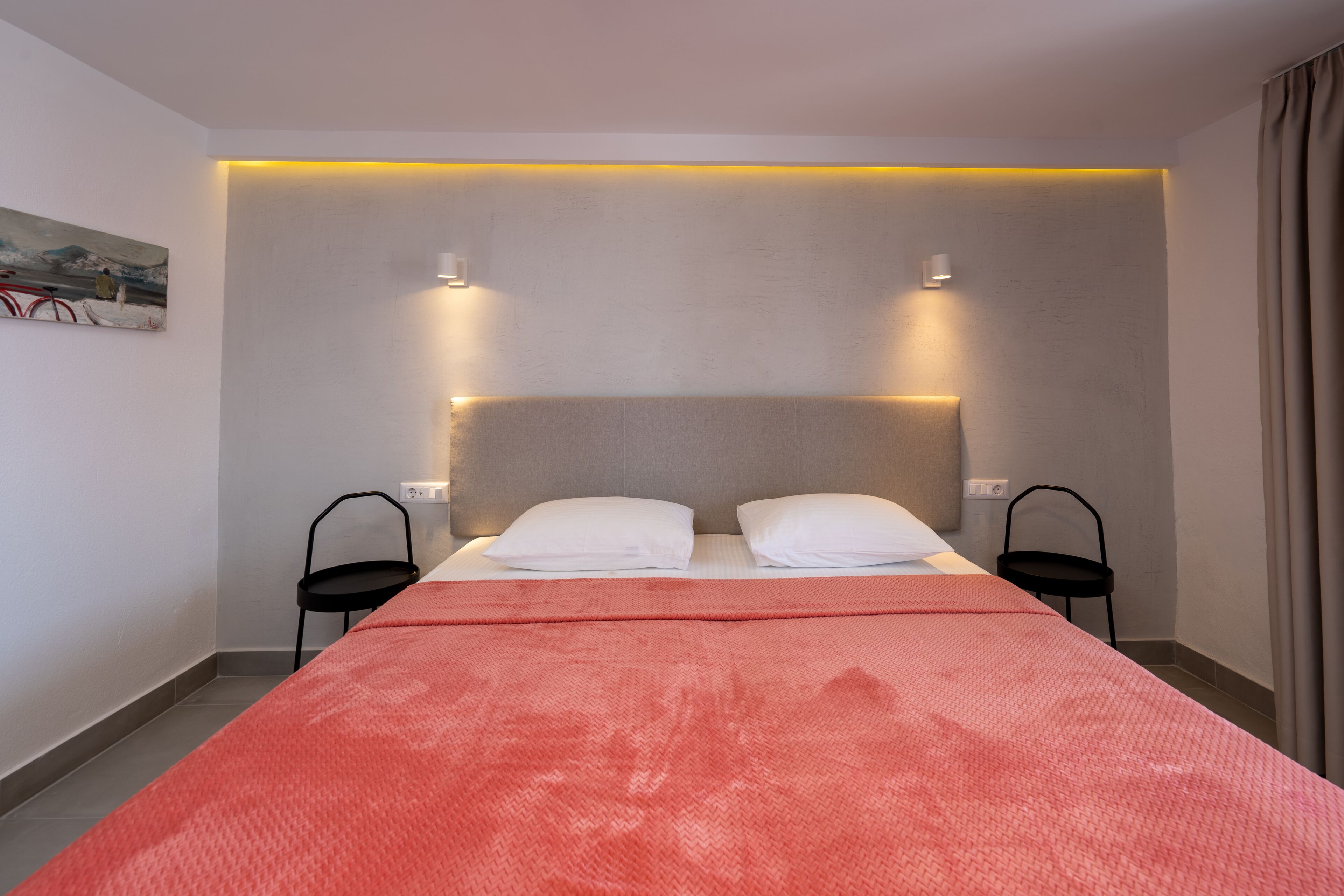 A hotel room with a bed, two bedside tables, a headboard with integrated lighting, a painting on the wall, and a window with curtains.