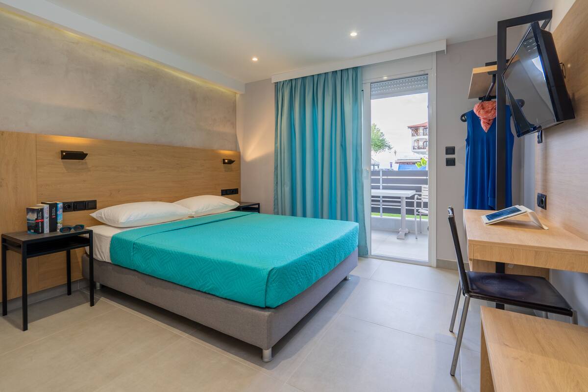 Quarto de hotel moderno com cama, escrivaninha, cadeira, TV e porta de vidro deslizante para o exterior.