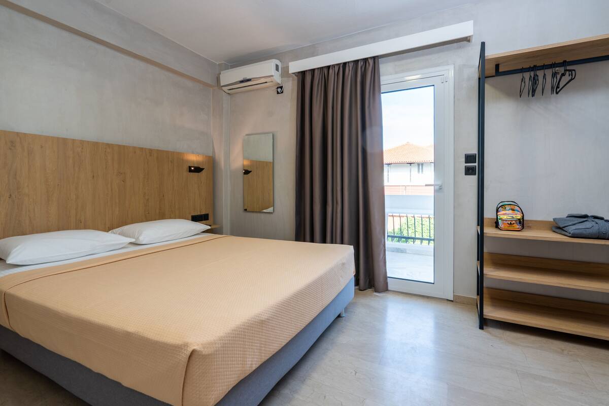 Quarto de hotel com cama, ar condicionado, espelho, janela com cortinas e prateleira com objetos.