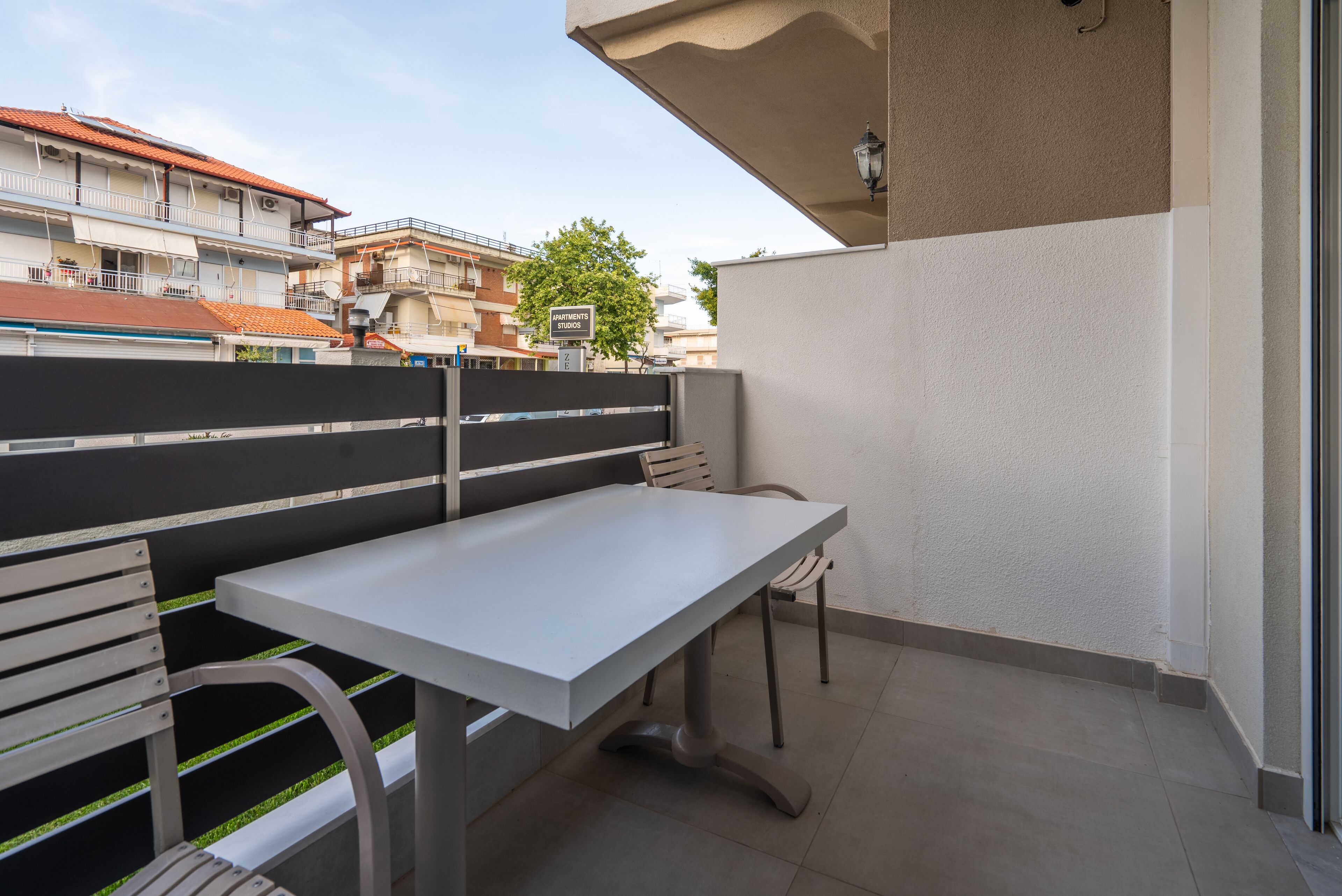 A balcony with a white table and chairs, a black and white striped fence, and a view of buildings and a tree in the background.