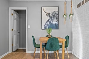 A dining area with a wooden table and green chairs, a framed artwork of a bull, and macrame hangings.