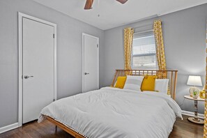 Une chambre à coucher avec un lit, deux portes, une fenêtre avec des rideaux, une table de chevet avec une lampe et un ventilateur de plafond.