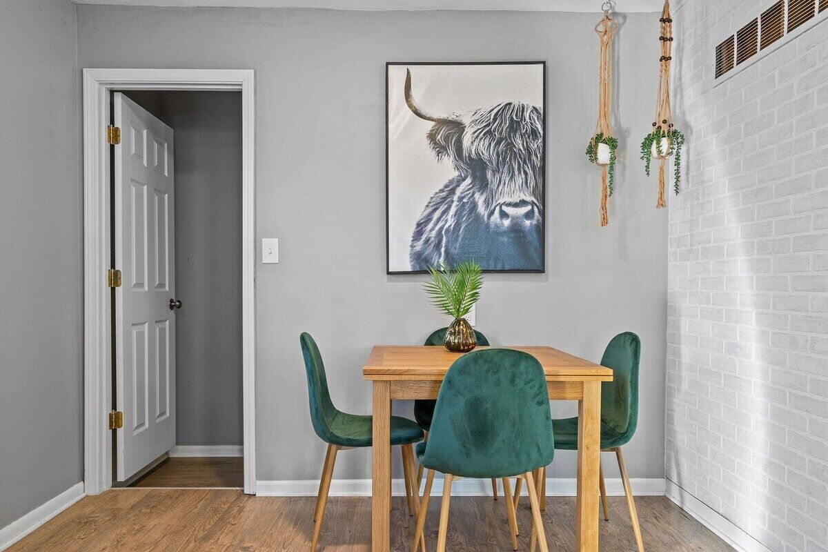 Ein Essbereich mit einem Holztisch und vier grünen Stühlen, ein gerahmtes Kunstwerk mit einem Stierkopf an der Wand und eine Tür, die in einen anderen Raum führt.