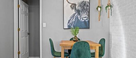 Ein Essbereich mit einem Holztisch und vier grünen Stühlen, ein gerahmtes Kunstwerk mit einem Stierkopf an der Wand und eine Tür, die in einen anderen Raum führt.