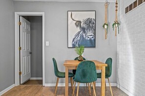 Un coin repas avec une table en bois et quatre chaises vertes, une œuvre d’art encadrée représentant la tête d’un taureau accrochée au mur, et une porte donnant sur une autre pièce.