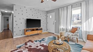 Ein modernes Wohnzimmer mit Flachbildfernseher, Deckenventilator, Wand mit Muster und großem Fenster mit Vorhängen.