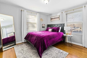 A bedroom with a bed, a round side table, a mirror, and a window with blinds.