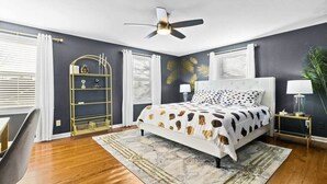 Un dormitorio con una cama, ventilador de techo, mesita de noche, lámpara, una silla y una estantería decorativa.