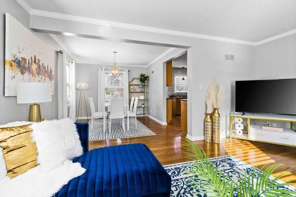 A modern living room with a blue sofa, a flat-screen TV, and a dining area with a chandelier.