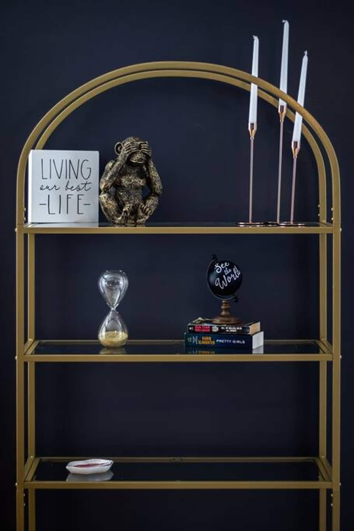 Una repisa dorada con un cartel que dice 'VIVIENDO nuestra mejor VIDA', una ampulleta de arena, libros y un globo terráqueo con la frase 'See the World'.