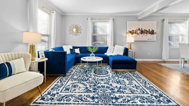 Una sala de estar con un sofá seccional azul, un sillón blanco, un tapete estampado y una obra de arte con vista a la ciudad en la pared.