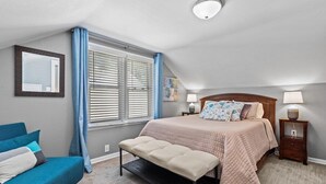 Ein Schlafzimmer mit einem Bett, Nachttischen, einer Bank, einem Fenster mit Jalousien und einer Deckenleuchte.