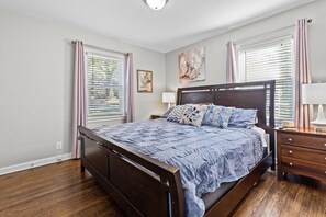 A bedroom with a wooden bed, bedside tables, a nightstand, and a window with blinds. - Jefferson St. Cottage - Large Home w/ Fenced Yard (Kansas City)
