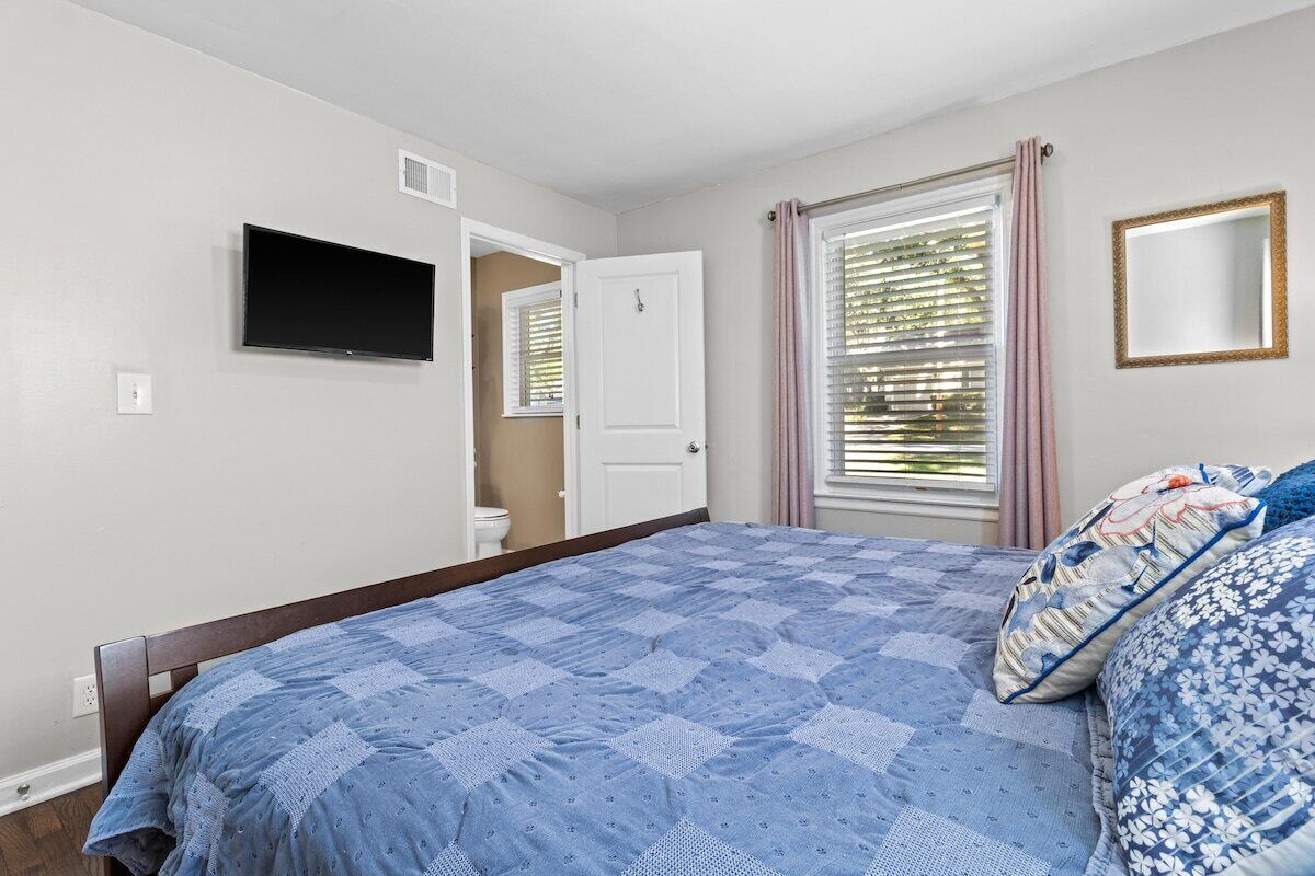 A bedroom with a bed, a television mounted on the wall, a window with blinds, and a door leading to another room.