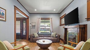 Ein Wohnzimmer mit braunem Ledersofa, zwei grünen Sesseln, einem Holztisch, einem Flachbildfernseher über einem Kamin und einem großen Fenster mit Vorhängen.
