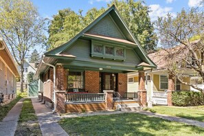 A two-story house with a green roof, brick foundation, and a front porch with a white railing.