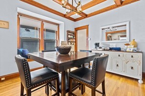 A dining area with a wooden table and chairs, a chandelier, and a kitchen area with cabinets and a mirror. - KC Nest - Walkable to Plaza + Parking + Streetcar (Kansas City)