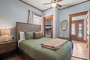 A bedroom with a bed, nightstand, lamp, ceiling fan, and a door leading to another room.