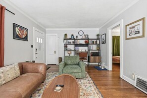 Un salon avec un canapé en cuir marron, un fauteuil vert et une table basse en bois. On y trouve une bibliothèque remplie de divers objets et une horloge au mur.