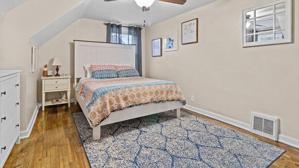 A bedroom with a bed, a ceiling fan, a window with curtains, a nightstand with a lamp, and a dresser. - Cottage Mins From Stadiums + Parking + Pet Welcome (Independence)