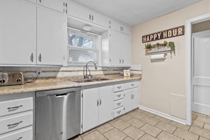 A kitchen with white cabinets, a stainless steel dishwasher, a window with blinds, and a 'HAPPY HOUR' sign on the wall.