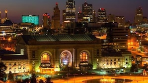 Vista urbana noturna com edifícios iluminados e um prédio histórico imponente com janelas em arco.