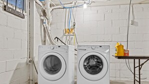 Two front-loading washing machines in a laundry room with white brick walls, visible plumbing, and a table with cleaning supplies. - Cottage Mins From Stadiums + Parking + Pet Welcome (Independence)