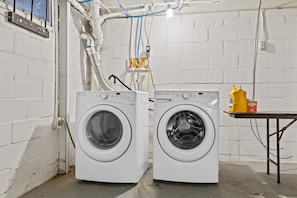 Zwei Waschmaschinen mit Frontlader in einem Waschraum mit weißen Ziegelwänden, freigelegter Sanitärinstallation und einem Tisch mit Reinigungsmitteln.
