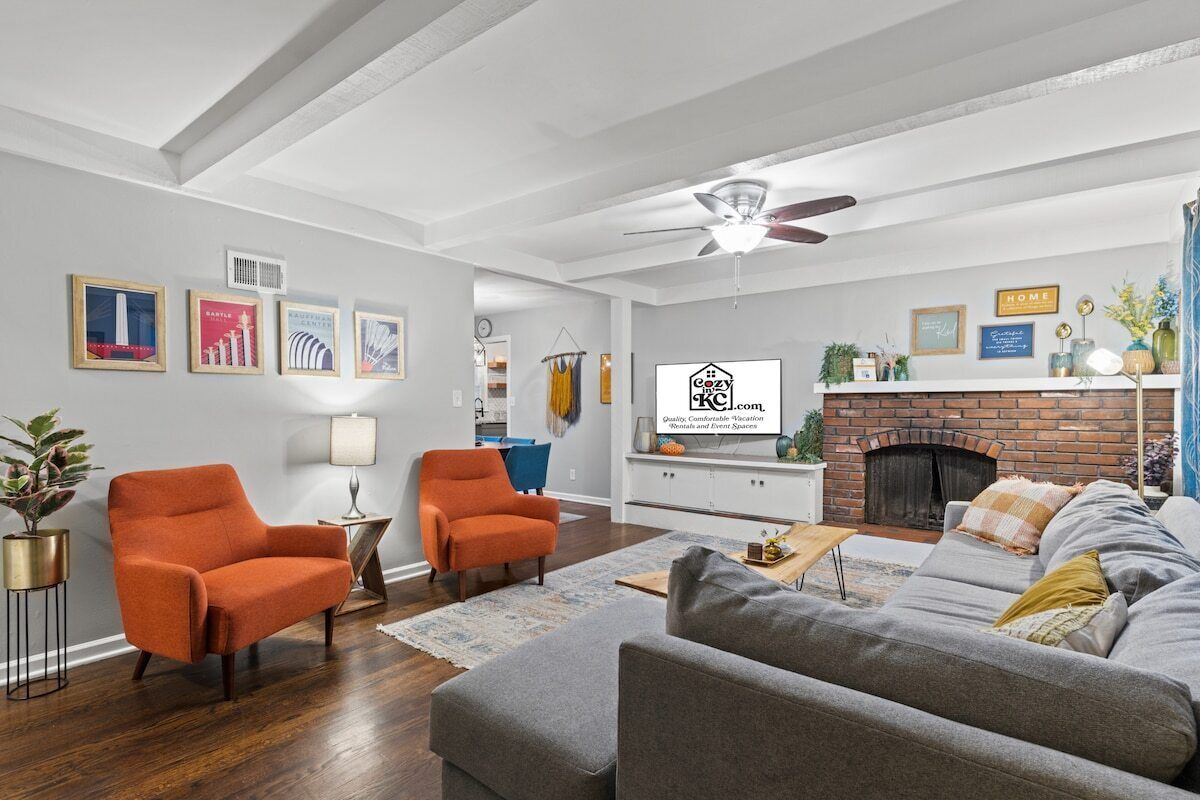 Un salon avec une cheminée, deux fauteuils orange, un canapé gris et un ventilateur de plafond.