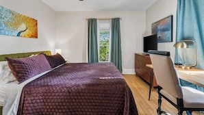 Quarto com cama, escrivaninha, cadeira, ventilador de teto e janela com cortinas.