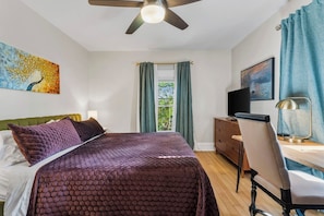 A bedroom with a bed, a desk, a chair, a ceiling fan, and a window with curtains.