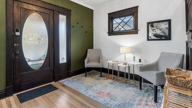 Une pièce avec une porte en bois sombre, un mur vert, une fenêtre, un lustre, deux chaises, une petite table et un tapis.
