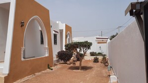 Una estrecha calle iluminada por el sol, con edificios blancos y anaranjados, arcos y macetas con plantas.