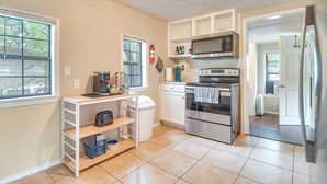 A compact kitchen with white cabinets, a microwave, an oven, and a refrigerator. There is a window with blinds, a shelf with kitchen items, and a trash can. - Half-Mi to Marina: Pet-Friendly Disney Home (Disney)