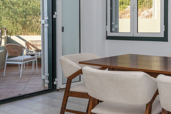 Una habitación con una mesa de madera y cuatro sillas, y un ventanal corredero que da a un área exterior con sillas de mimbre.