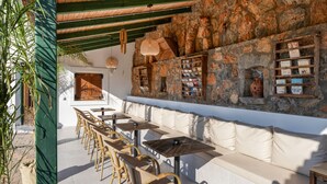 Un espace extérieur aménagé avec des chaises et des tables en osier, un mur en pierre agrémenté d’étagères et une porte en bois.